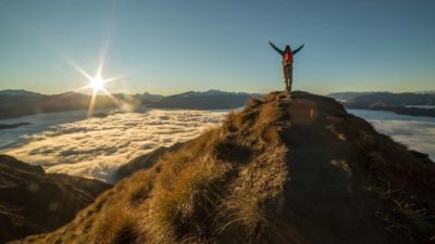 a person stands on top of a mountain with hands raised above their head gazing on an amazing sunrise over the landscape and above the clouds.