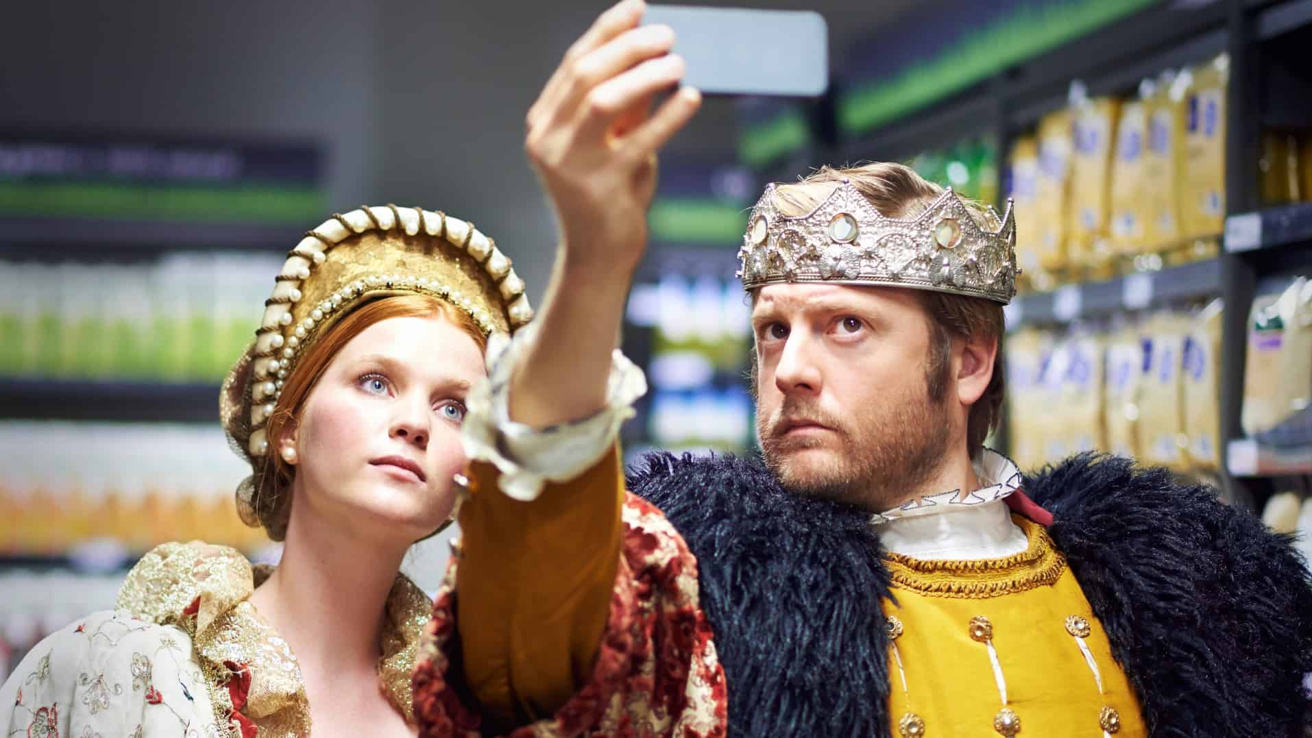 a man and a woman in historical costume, Henry the eitghth era, post for a selfie with the man holding a phone above their heads while the two pose with serious faces as seen in historical portraits of people.