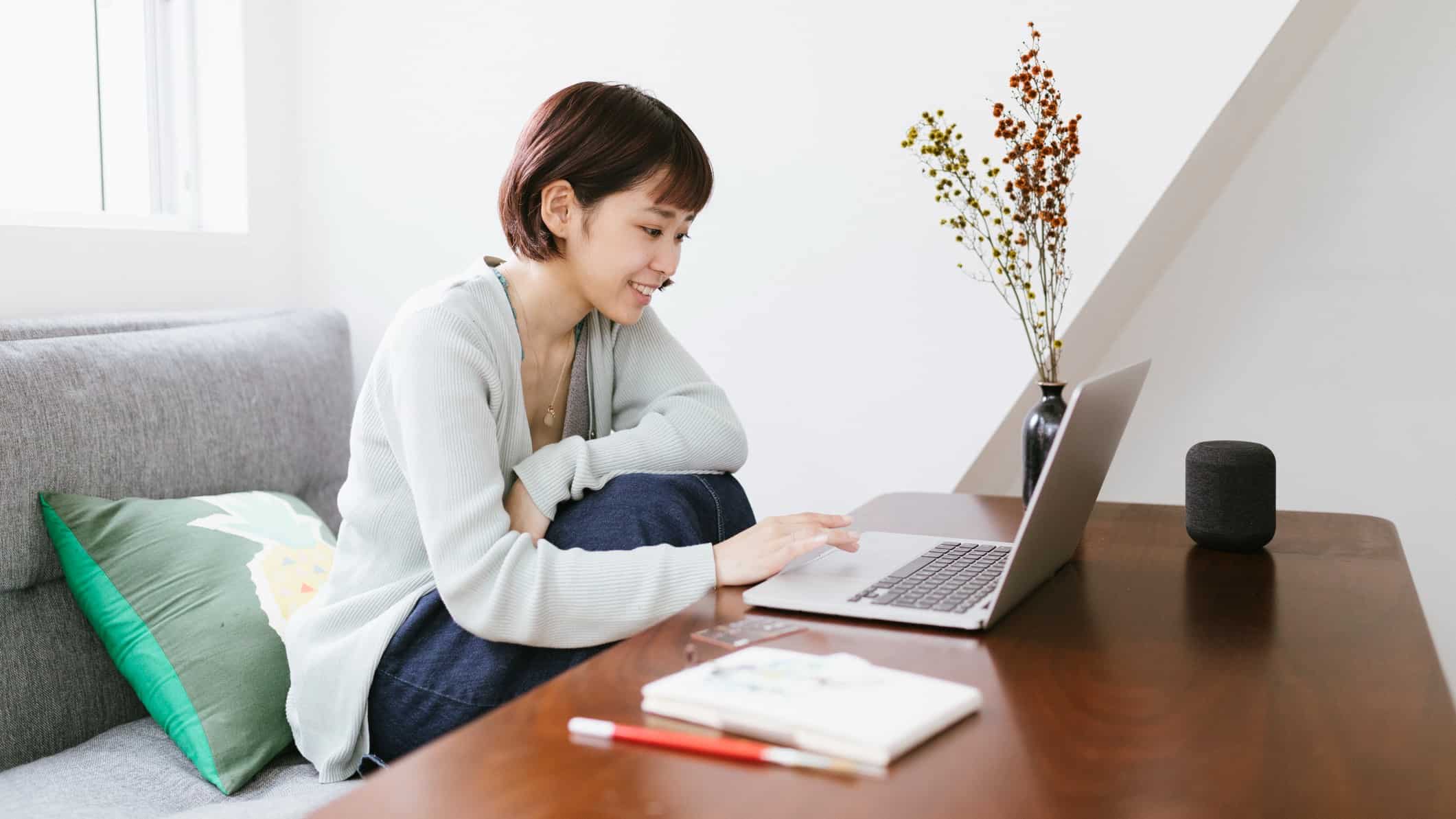 a woman sits in a quiet home nook with her laptop computer and a notepad and pen on the table next to her as she smiles at information on the screen.