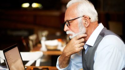 an elderly man holds his chin in concern as he looks at his computer screen.