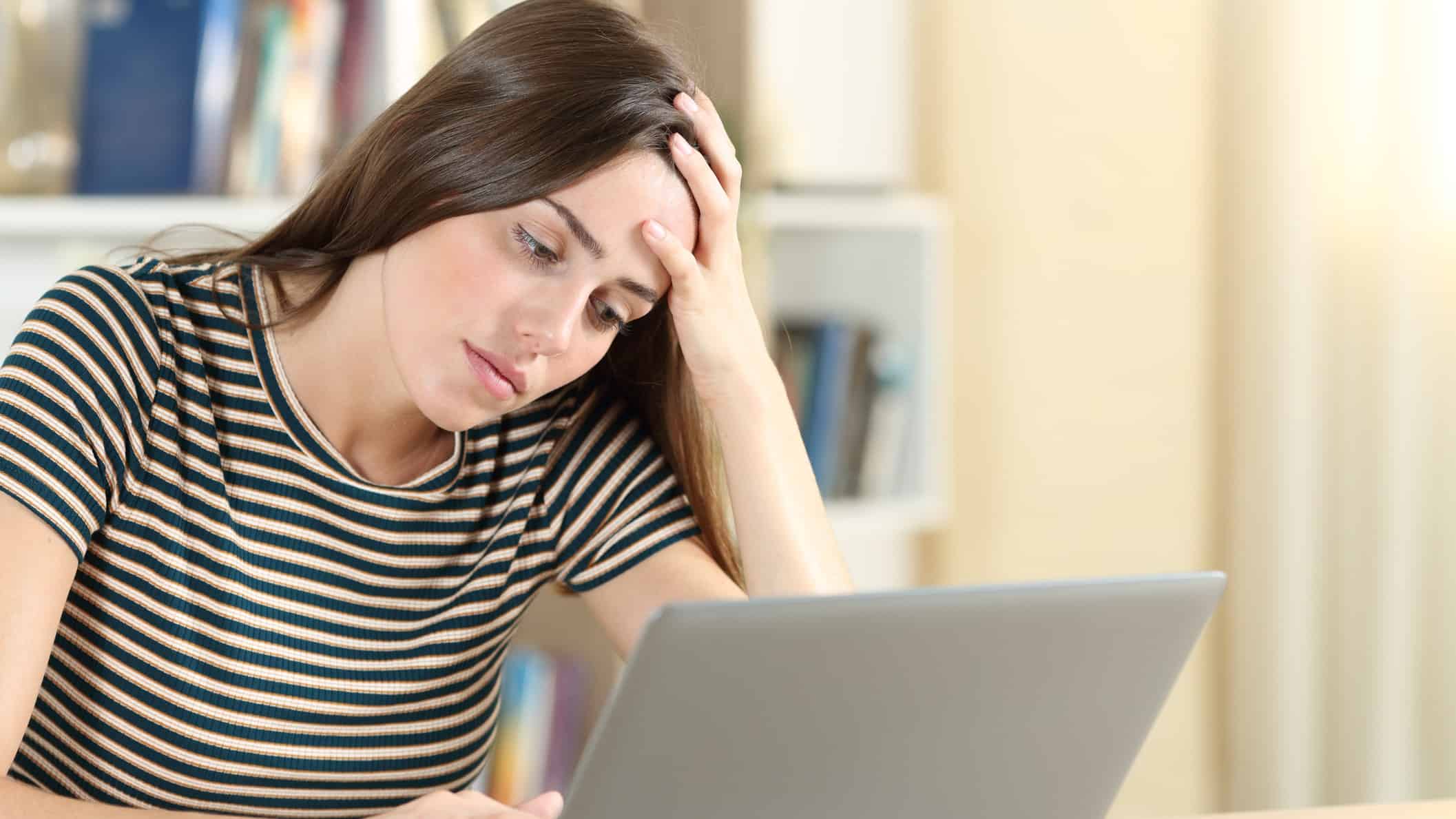 a woman looks exhausted and overwhelmed as she slumps forward into her hand while looking at her laptop screen.