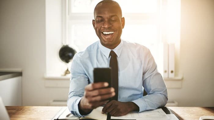 a man sits at his desk wearing a business shirt and tie and has a hearty laugh at something on his mobile phone.