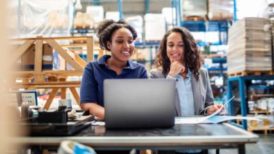 two women stand at a computer smiling in a large factory with high shelves piled with goods, as though working in logistics.