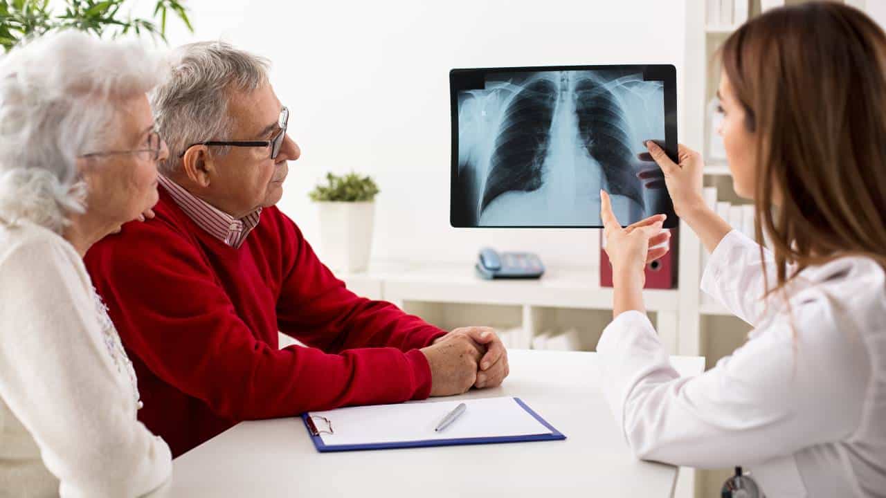 A doctor and an elderly couple sit at a desk and look at a lung scan uploaded using Alcidion software as the Alcidion share price falls