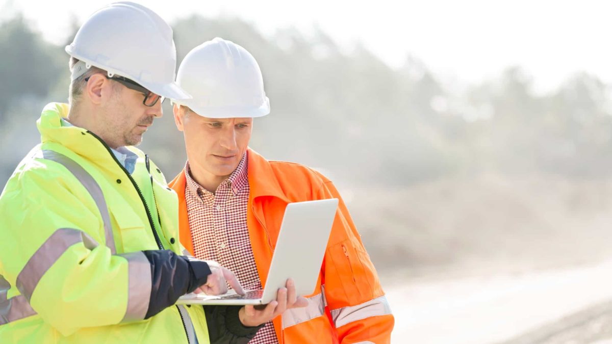 two men in hard hats and high visibility jackets look together at a laptop screen that one of the men in holding at a mine site.