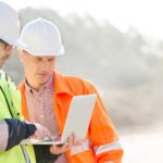 Two men in hard hats and high visibility jackets look together at a laptop screen at a mine site.