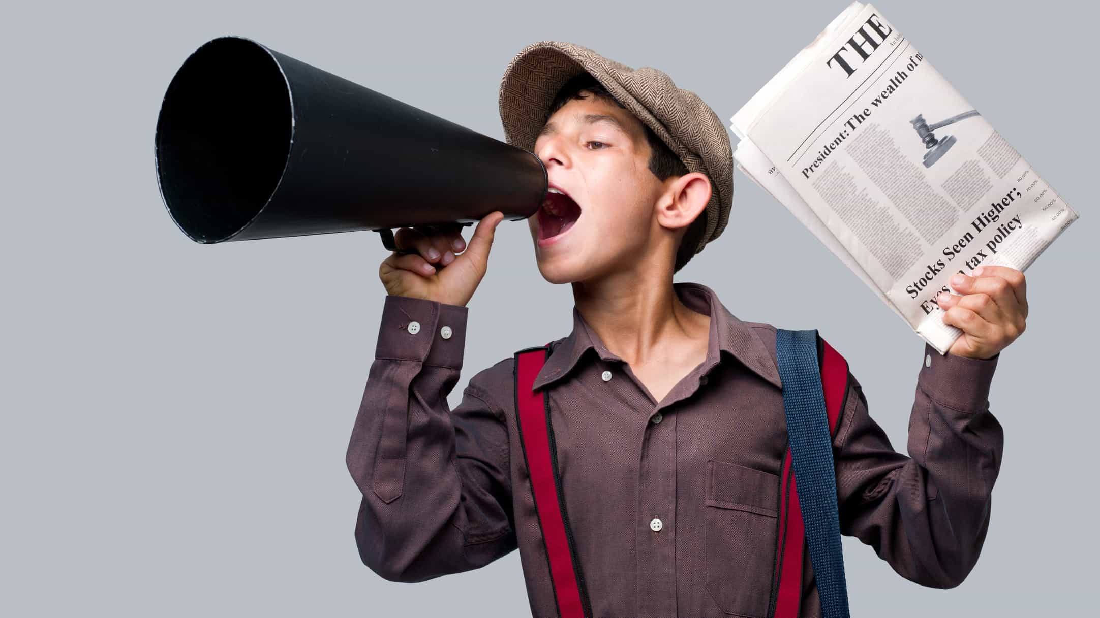 a newsboy wearing historical costume of peaked cap and braces yells into an old fashioned megaphone while holding a newspaper in one hand, a so-called newsboy of previous eras when newsboys sold newspapers on street corners.