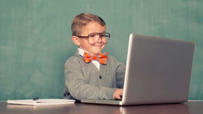 A young boy dressed in an old man-style cardigan with business shirt and bow tied wearing big spectacles smiles to himself as he sits at a laptop computer at a desk with hands on keys.