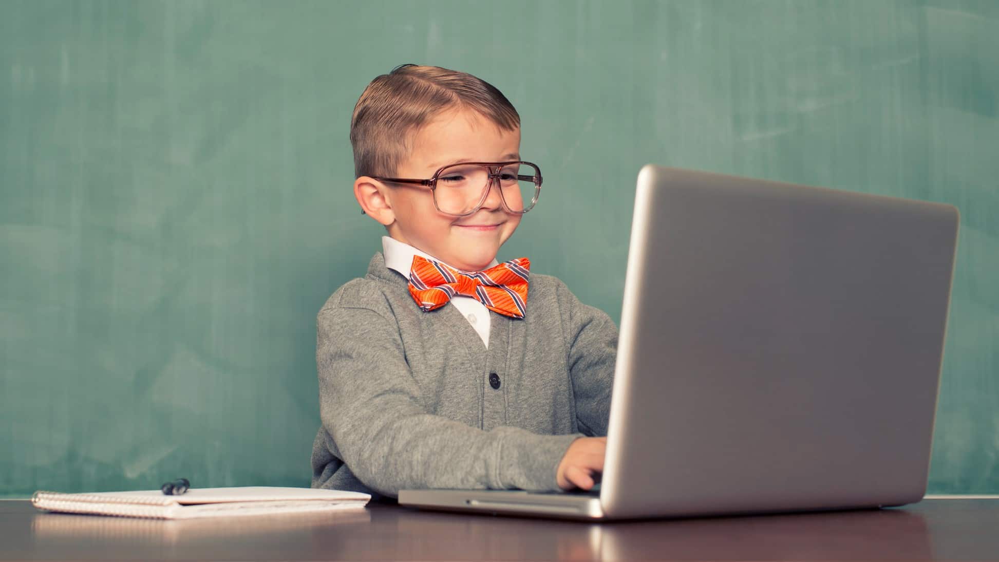 A young boy dressed in an old man-style cardigan with business shirt and bow tied wearing big spectacles smiles to himself as he sits at a laptop computer at a desk with hands on keys.