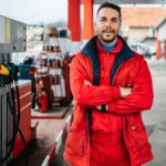 A service station attendant crosses his arms and smiles towards the camera with a backdrop of petrol bowsers and a drive-through facility.