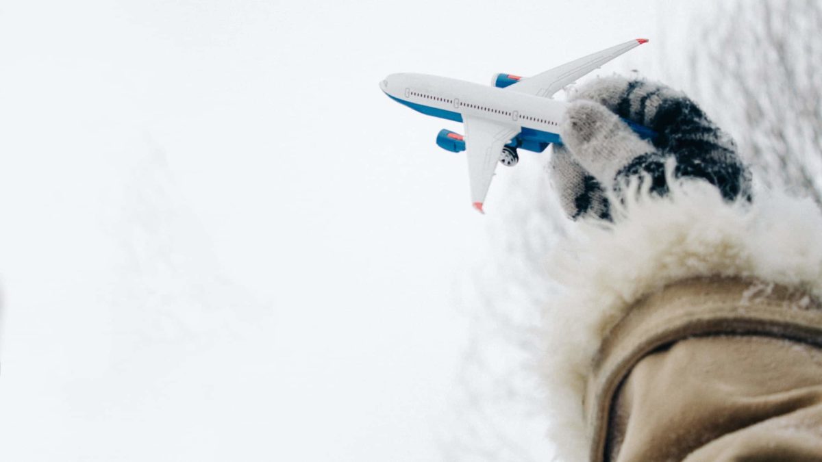 a gloved hand with a fur lined jacket attached holds a small toy aeroplane against a frozen white, icy backdrop.