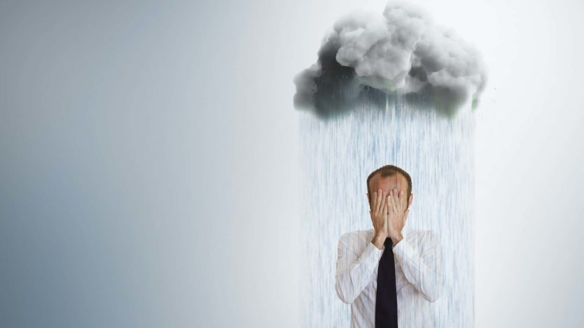 A man in a business suit covers his face with his hands as he stands under a storm cloud emitting heavy rain on top of him.