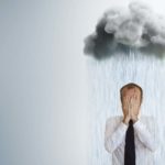 A man in a business suit covers his face with his hands as he stands under a storm cloud emitting heavy rain on top of him.