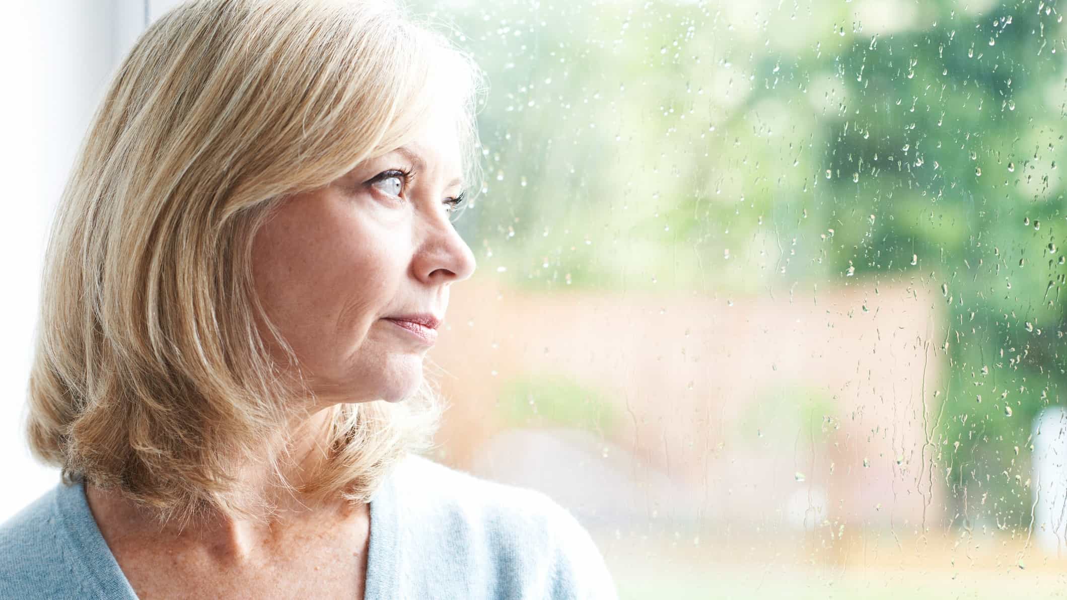 a woman looks out a window splattered with rain with a pensive look on her face.