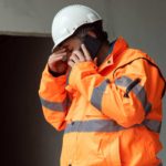 a sad looking engineer or miner wearing a high visibility jacket and a hard hat stands alone with his head bowed and hand to his forehead as he speaks on a mobile telephone out front of what appears to be an on site work shed.