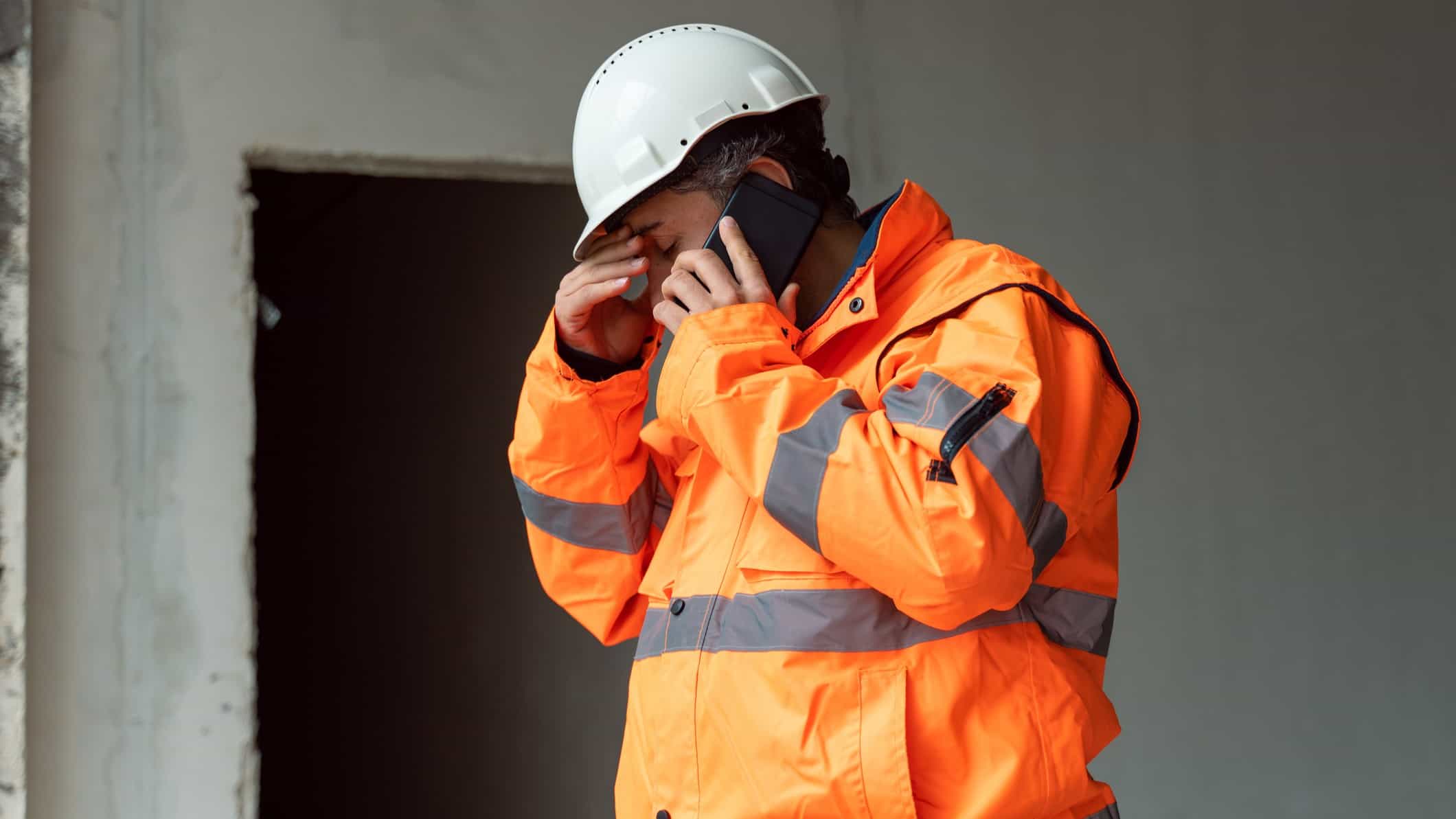 a sad looking engineer or miner wearing a high visibility jacket and a hard hat stands alone with his head bowed and hand to his forehead as he speaks on a mobile telephone out front of what appears to be an on site work shed.