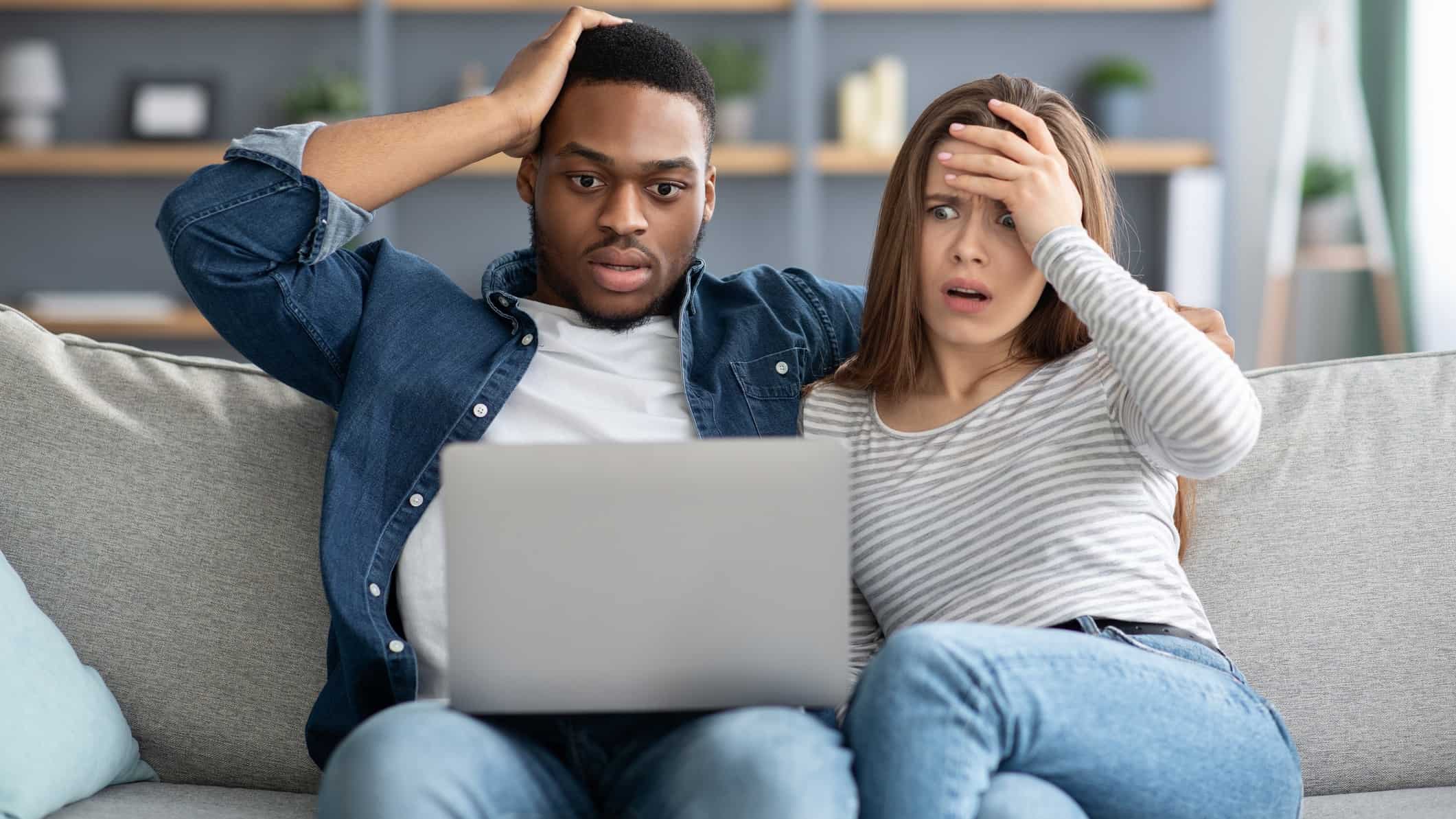 a couple sits on a sofa, each clutching their heads in horror and disbelief, while looking at the computer screen balanced on the lap of the man.