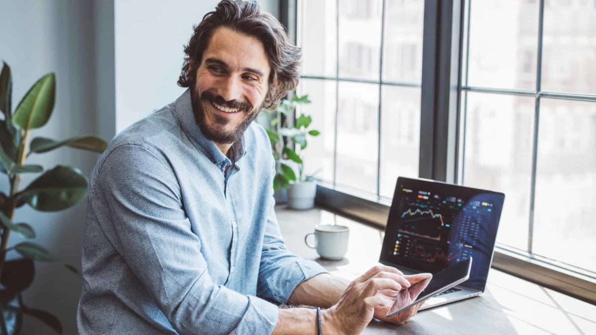 Smiling man sits in front of a graph on computer while using his mobile phone.