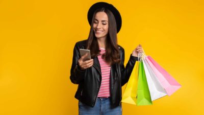 a young woman looks happily at her phone in one hand with a selection of shopping bags in her other hand.