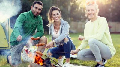 Three people outdoors cooking sausages over a fire.