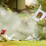 Two children dressed as space travellers in white suits look on at the smoking wreckage of their tin foil covered carboard rocket in their backyard with one child pulling the other away from the crash site.