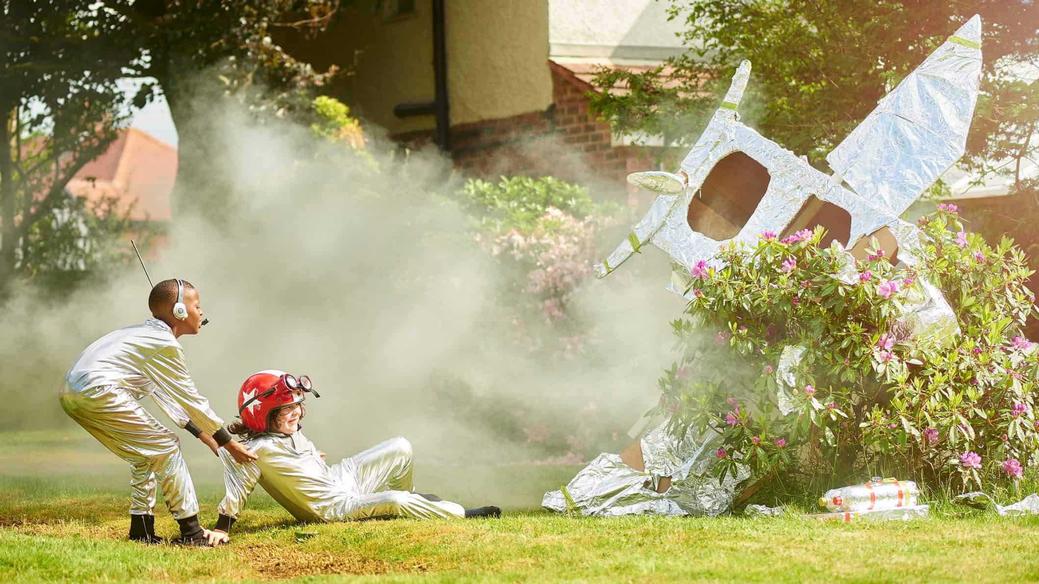 Two children dressed as spacemen in white suits look on at the smoking wreckage of their tin foil covered carboard rocket in their backyard with one child pulling the other away from the crash site.