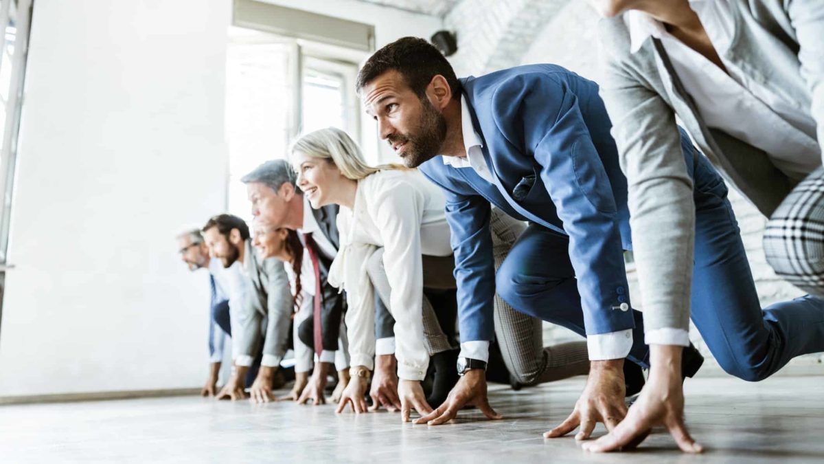 a group of seven businesspeople take to the floor in starter block positions as though they are set to compete in a running race in an office environment.