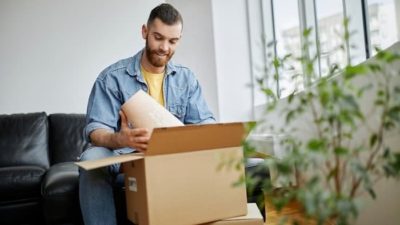 a man smiles widely as he opens a large brown box and examines the contents in his home.