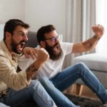 two men raise their fists and shout with their mouths wide open on a sofa as though they are watching sport or something stirring on a television that is out of picture.