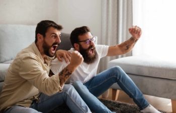 two men raise their fists and shout with their mouths wide open on a sofa as though they are watching sport or something stirring on a television that is out of picture.