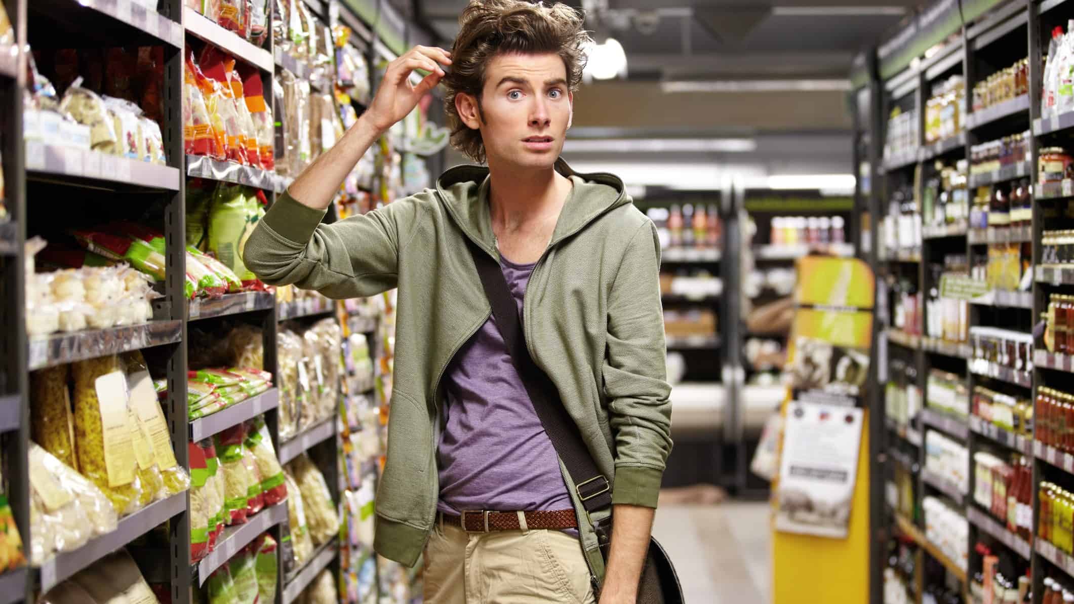 a man looks a little perplexed as he holds his hand to his head as if thinking about something as he stands in the aisle of a supermarket.