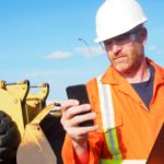 a mine worker holds his phone in one hand and a tablet in the other as he stands in front of heavy machinery at a mine site.