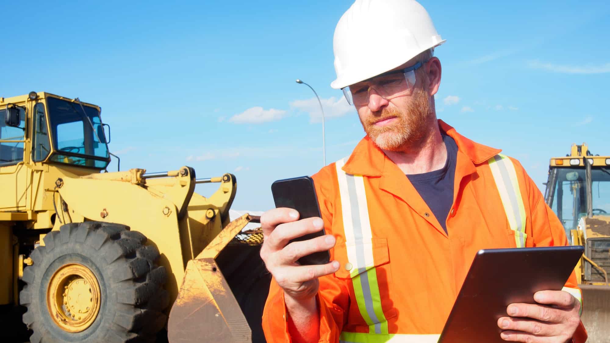 a mine worker holds his phone in one hand and a tablet in the other as he stands in front of heavy machinery at a mine site.
