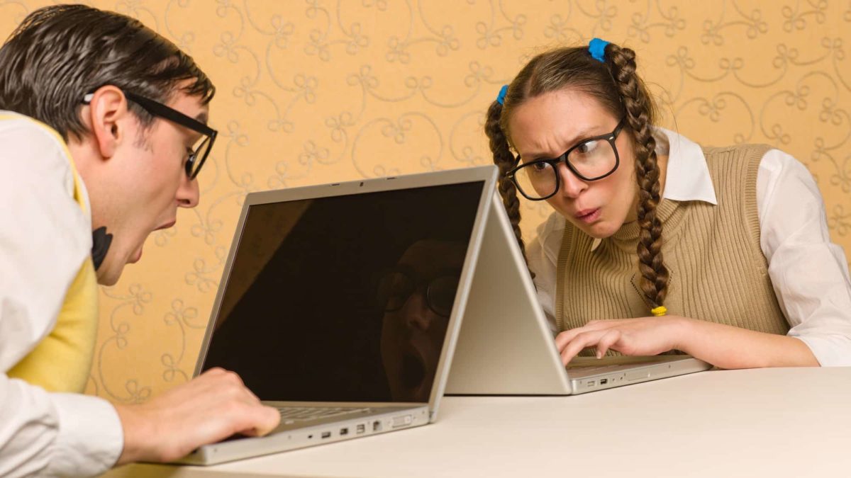 two computer geeks sit across from each other with their laptop computers touching as they look confused and confounded by what they are seeing on their screens.