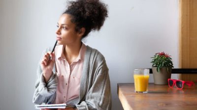 A woman sits at a table with notebook on lap and pen in hand as she gazes off to the side with the pen resting on the side of her face as though she is thinking and contemplating while a glass of orange juice and a pair of red sunglasses rests on the table beside her.