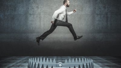 a man in a business shirt and tie takes a wide leap over a large steel trap with jagged teeth that is place directly underneath him.