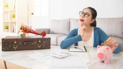 A woman ponders a question as she puts money into a piggy bank with a model plane and suitcase nearby.