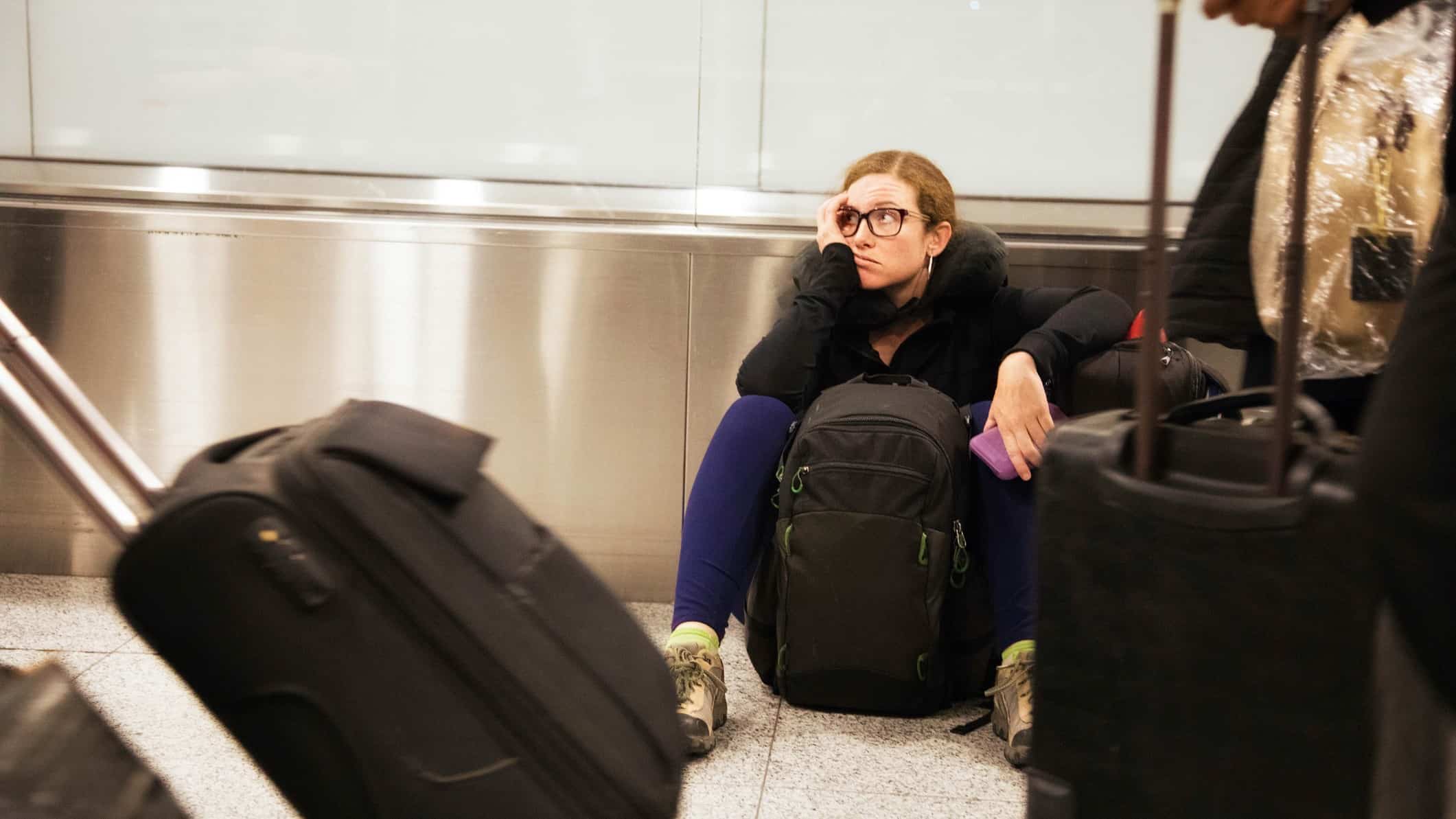 A traveller sits slumped on the floor of an airport with ehr suitcase looking fed-up as other travellers walk past.