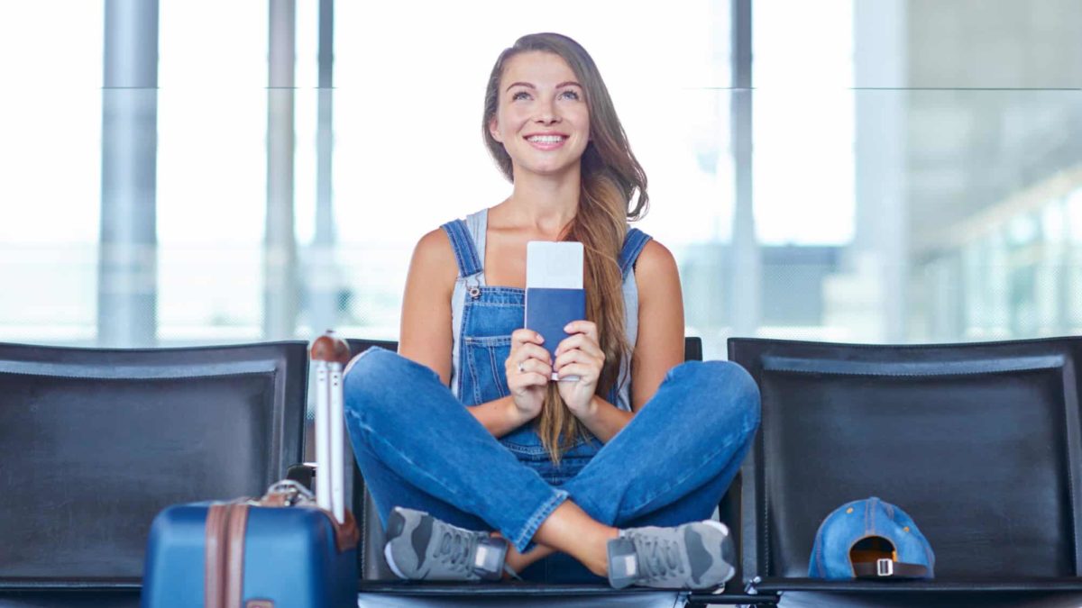A woman sits crossed legged on seats at an airport holding her ticket and smiling.