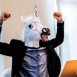 A man with a unicorn mask sits at desk and cheers.