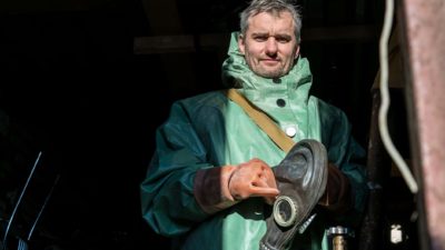 a uranium plant worker in full protective gear removes his head covering and holds it in his hand as he smiles slightly to have his picture taken.