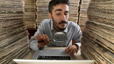 a man surrounded by huge piles of paper looks through a magnifying glass at his computer screen.