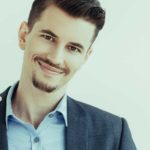 A young man wearing an open necked shirt and a stylish coat raises a glass of champagne as he smiles.