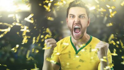 a man in a green and gold Australian athletic kit roars ecstatically with a wide open mouth while his hands are clenched and raised as a shower of gold confetti falls in the sky around him.
