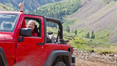 A man in a four wheel drive vehicle lifts an arm and gives a thumbs up in the air as he traverses rugged mountain style terrain with a green valley and rocky hills in the background.