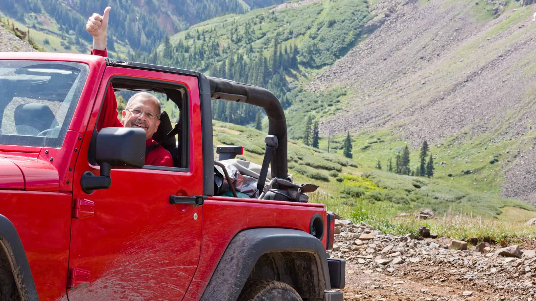 A man in a four wheel drive vehicle lifts an arm and gives a thumbs up in the air as he traverses rugged mountain style terrain with a green valley and rocky hills in the background.