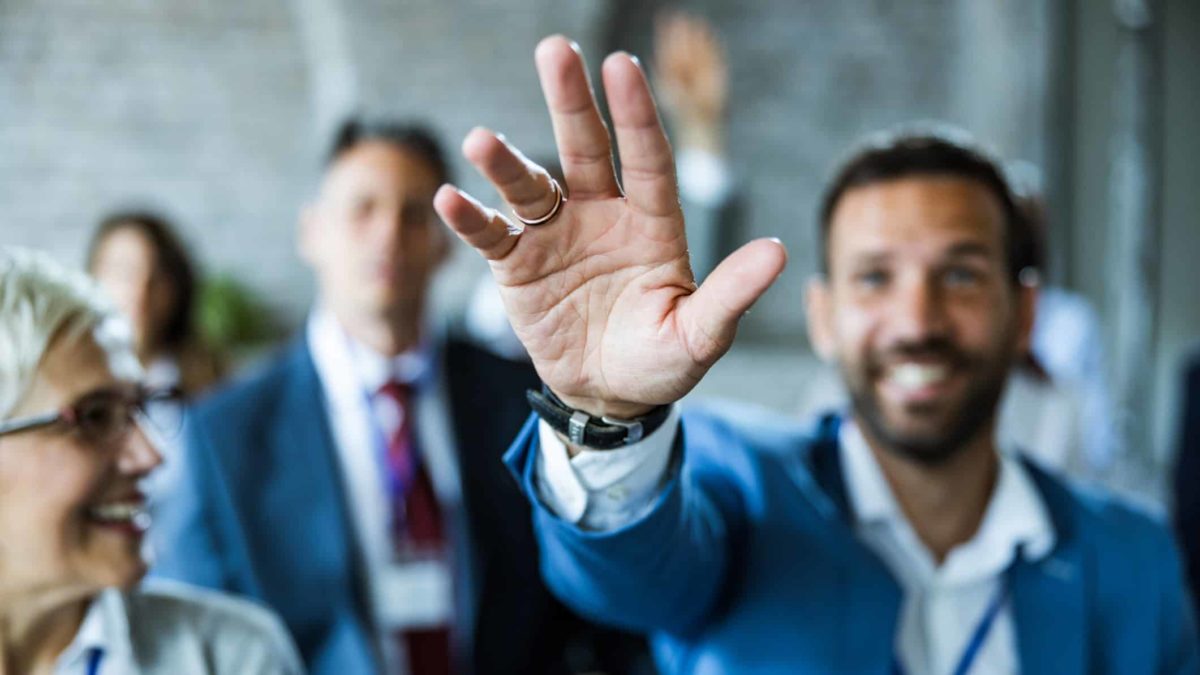 A group of people in suits watch as a man puts his hand up to take the opportunity.