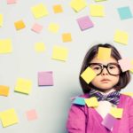 A young girl wearing glasses stares without smiling with lots of post-it notes stuck all over the wall behind her and all over her face.