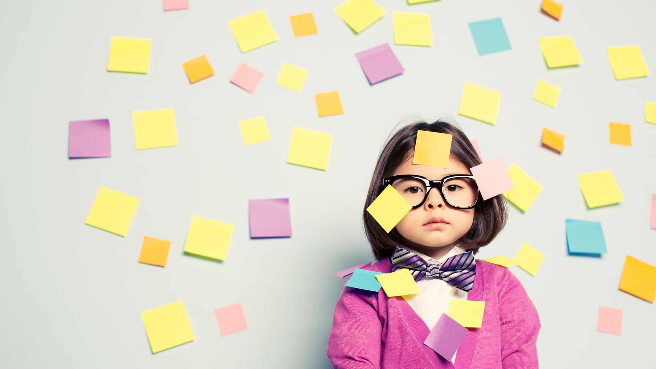 A young girl wearing glasses stares without smiling with lots of post-it notes stuck all over the wall behind her and all over her face.