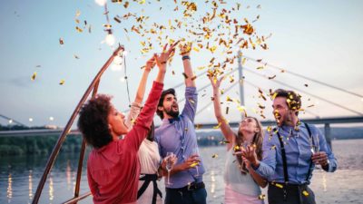 A group of friends throw gold confetti in the air in celebration as they sail on a boat on a river.